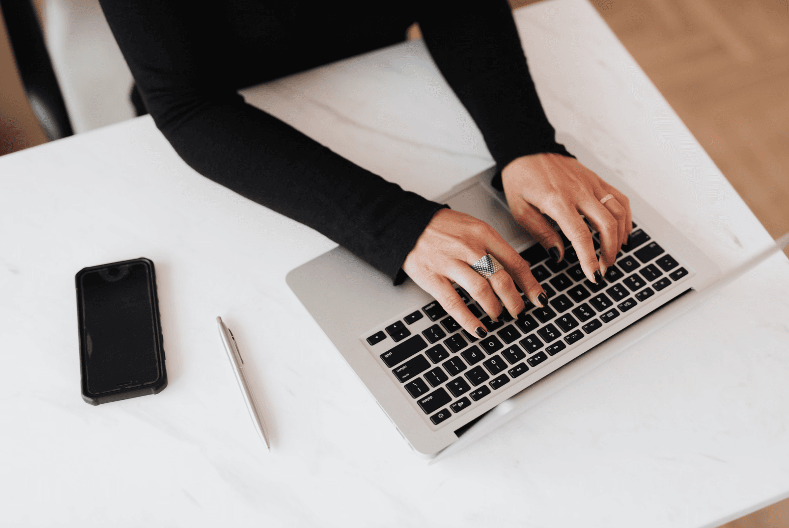 Hands typing on a laptop beside a phone and a pen on a white desk